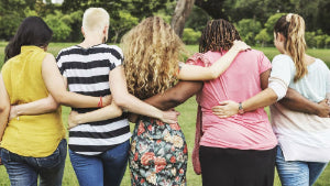 Five women standing outdoors in a park with their arms around each other, seen from behind. They are wearing different casual outfits in yellow, black and white stripes, floral print, pink, and light blue.